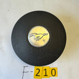 A black textured Mustang steering wheel center cap with the silver running horse emblem, placed on a light-colored surface with yellow numbered stickers and handwritten notes nearby.