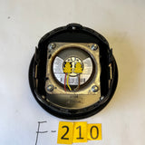 A circular black plastic housing containing a metallic circular component with yellow wire leads, photographed on a white tabletop with yellow paper labels reading 'F-210' below it.